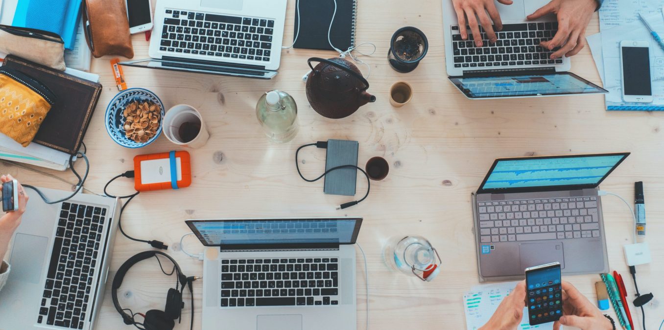 An overhead view of a busy wooden communal table where several people are working together. The desk is cluttered with multiple laptops showing data charts, smartphones, headphones, notebooks, and coffee cups, capturing a high-energy collaborative workspace.