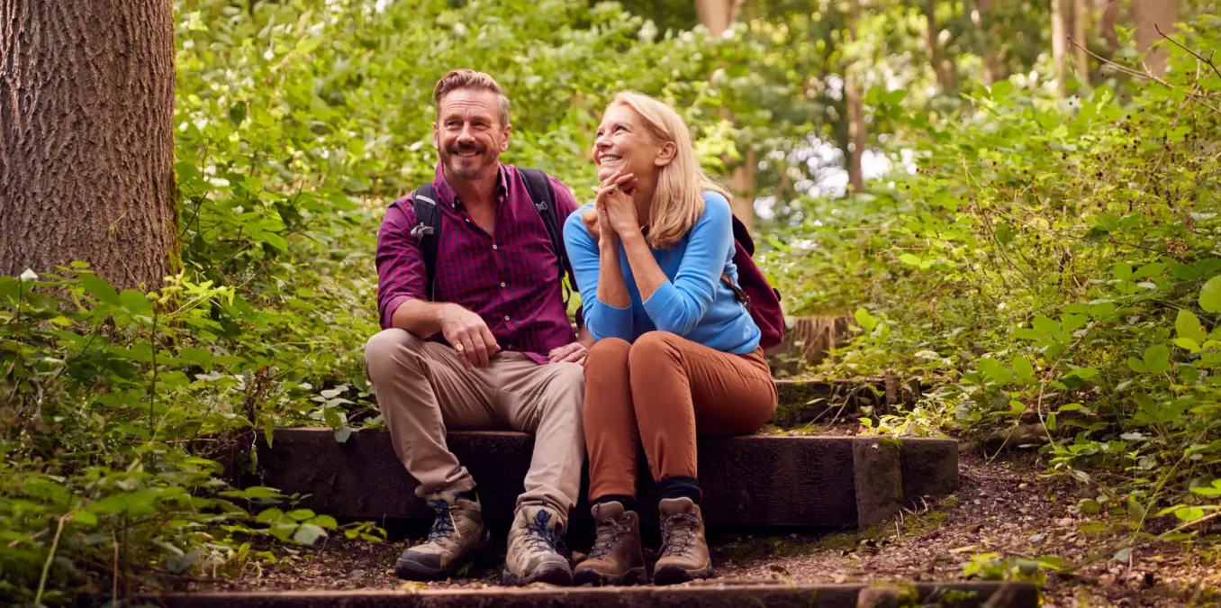 A smiling middle-aged man and woman taking a break during a hike, sitting together on rustic wooden stairs in a lush, sunlit green forest. The man wears a red plaid shirt and backpack, while the woman wears a blue long-sleeve shirt and looks at him admiringly. Both are wearing sturdy hiking boots.