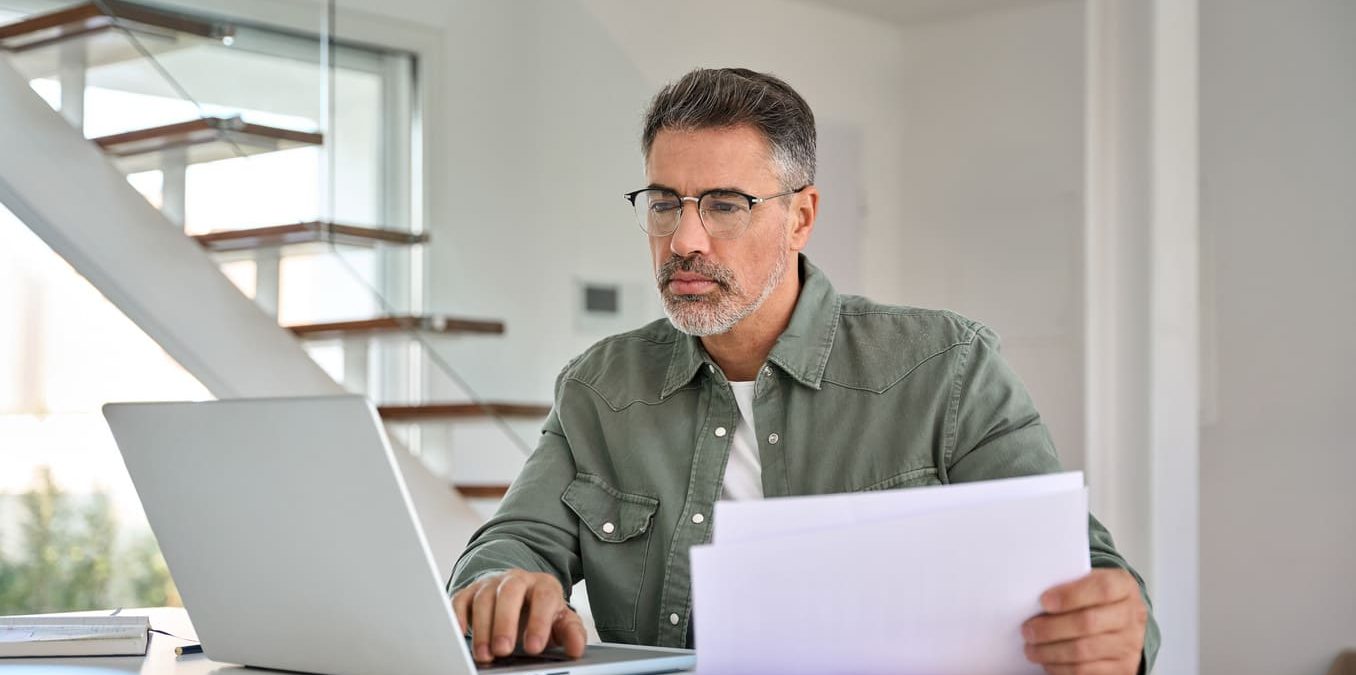 A middle-aged man with glasses sitting at a table, focused on his laptop while holding several sheets of paper in his home office