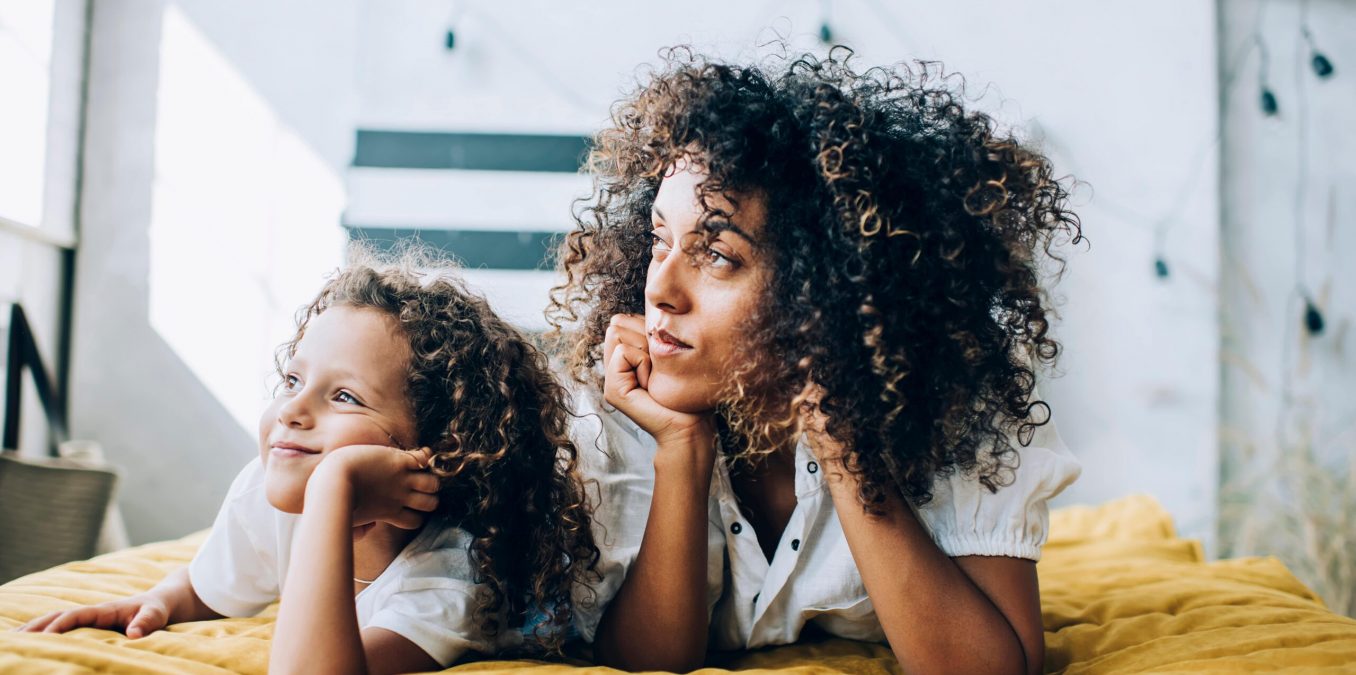 A woman and a young girl, both with voluminous curly hair, lying on their stomachs on a yellow textured bedspread. They are both resting their chins on their hands and looking off-camera with thoughtful, happy expressions in a bright, modern room.