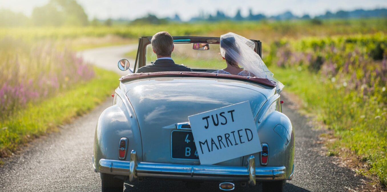 A rearview shot of a newlywed couple driving a vintage light blue convertible down a winding country road. A "Just Married" sign is attached to the back of the car, and the bride's white veil flutters in the wind as they drive through a sunny landscape.