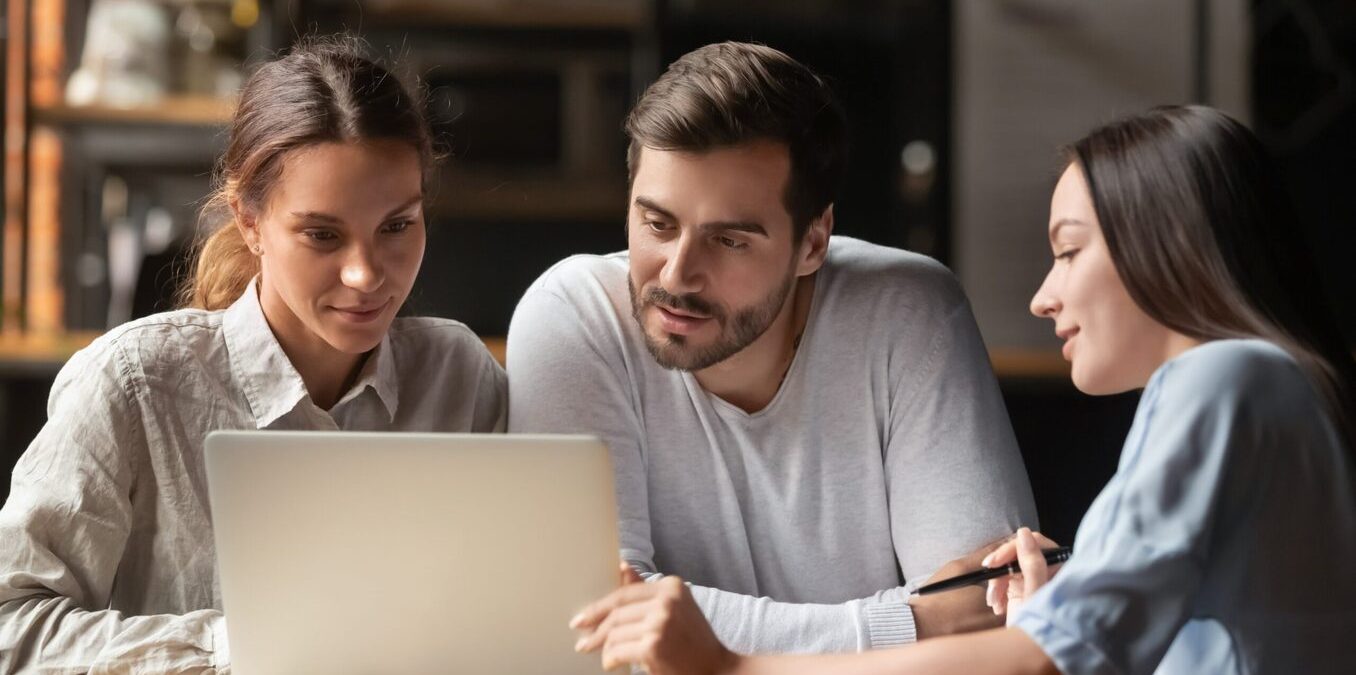 A professional woman in a light blue shirt points to a laptop screen while explaining information to a young couple. The man and woman lean in with focused expressions as they review the details together at a table in a modern office or cafe setting.