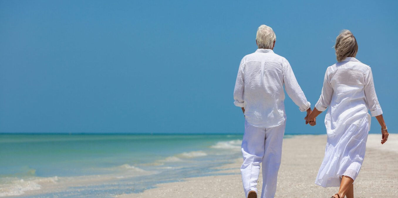 Rear view of happy senior man and woman couple walking and holding hands on a deserted tropical beach with bright clear blue sky