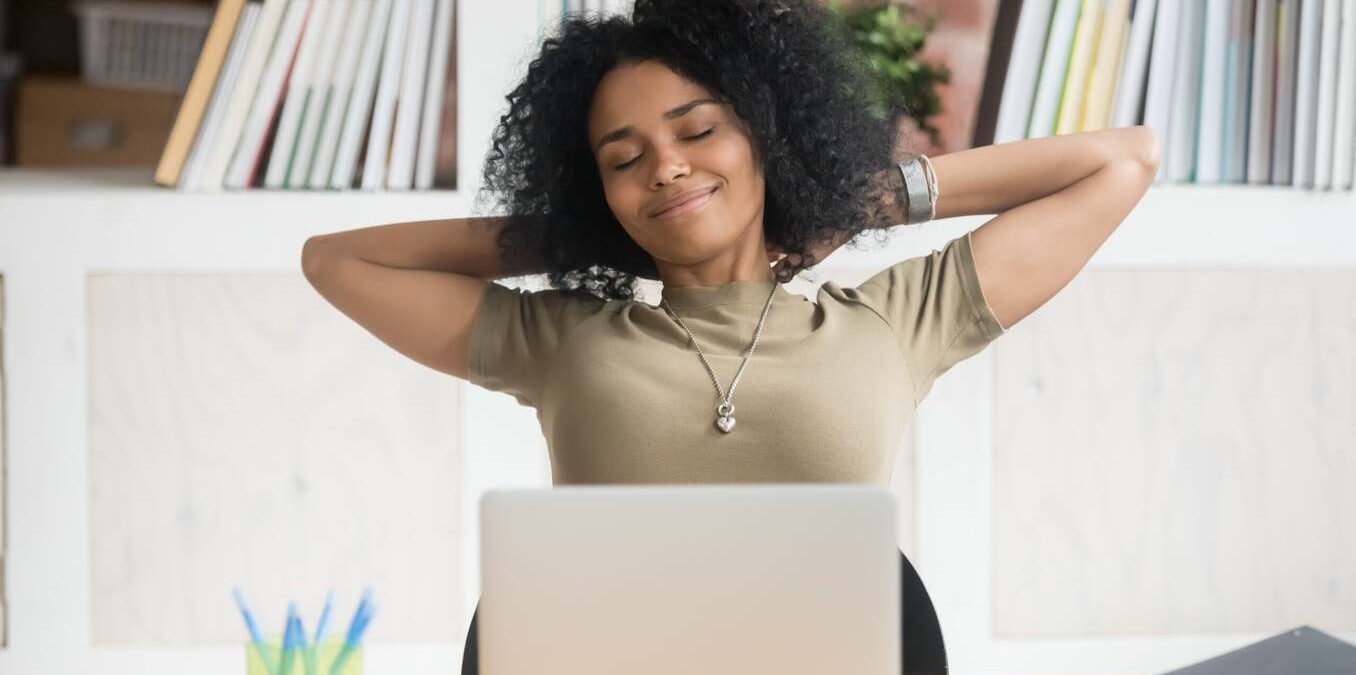 A woman sitting at a desk with a laptop, smiling with her eyes closed and hands behind her head as she stretches and takes a break