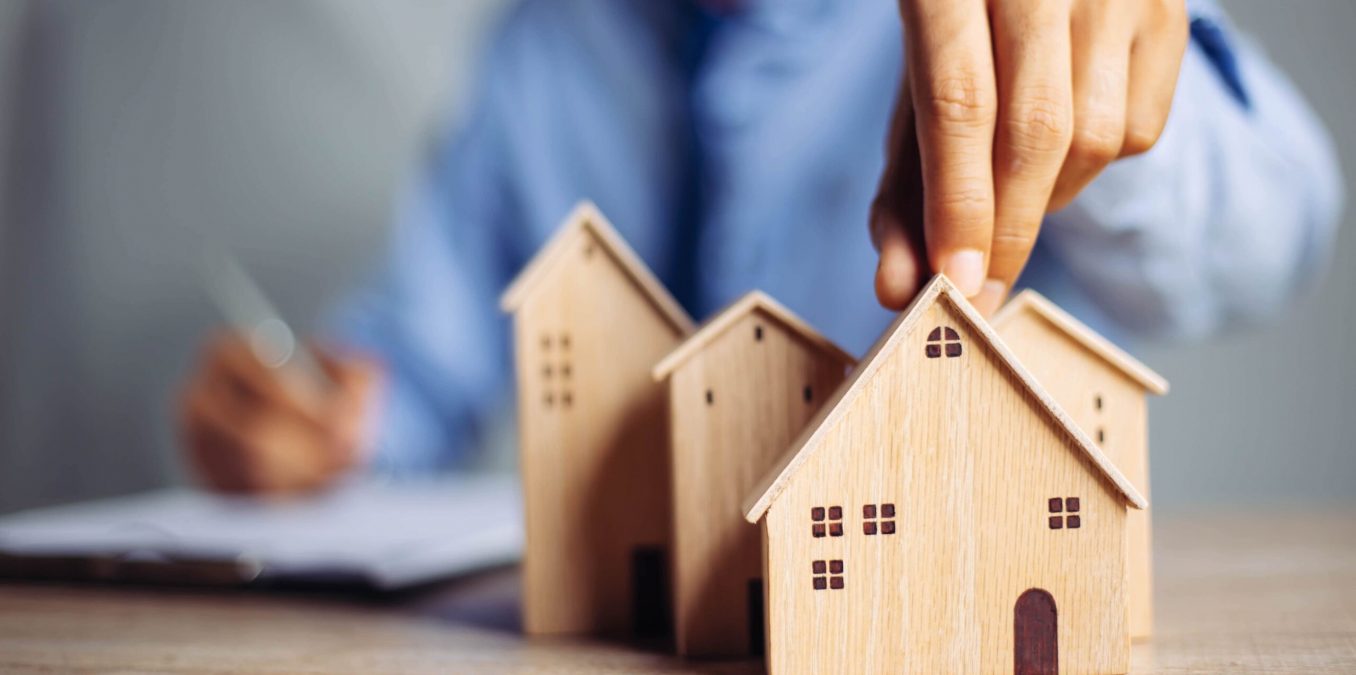A close-up of a person’s hand placing a small wooden house model among a cluster of other wooden houses on a desk. In the blurred background, a person in a blue shirt is writing on a clipboard, symbolizing real estate planning, home buying, or property investment.