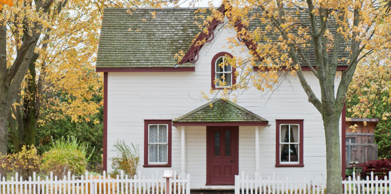 A charming two-story white historic farmhouse with a dark red trim and a gabled roof, framed by autumn trees with yellow leaves. A white picket fence runs across the foreground, and a small porch covers the front door, creating a classic rustic aesthetic.