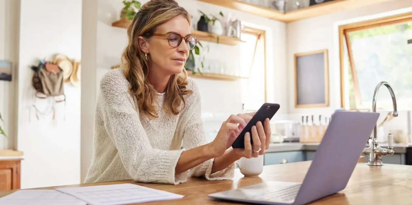 A mature woman wearing glasses sitting in a brightly lit kitchen, using her smartphone while working on a laptop with financial documents nearby to review Medicare IRMAA costs.