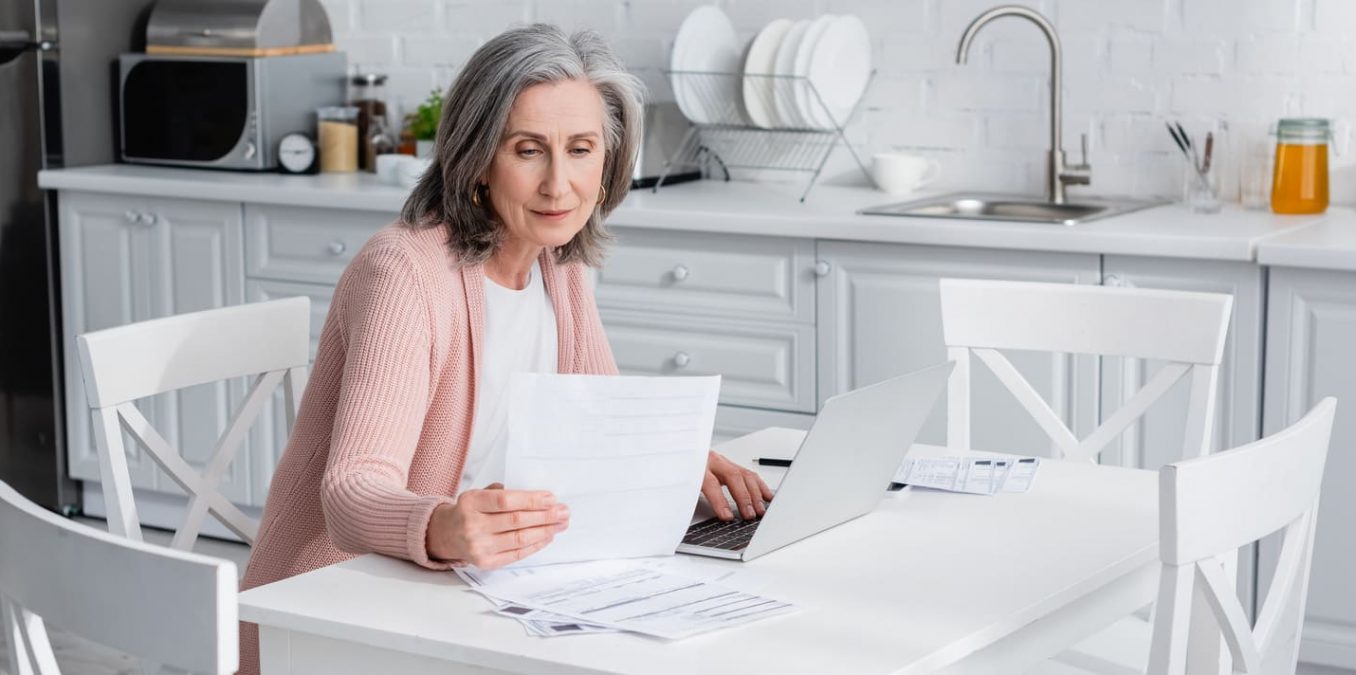 A senior woman sitting at a kitchen table reviewing financial paperwork and investment documents while using a laptop.