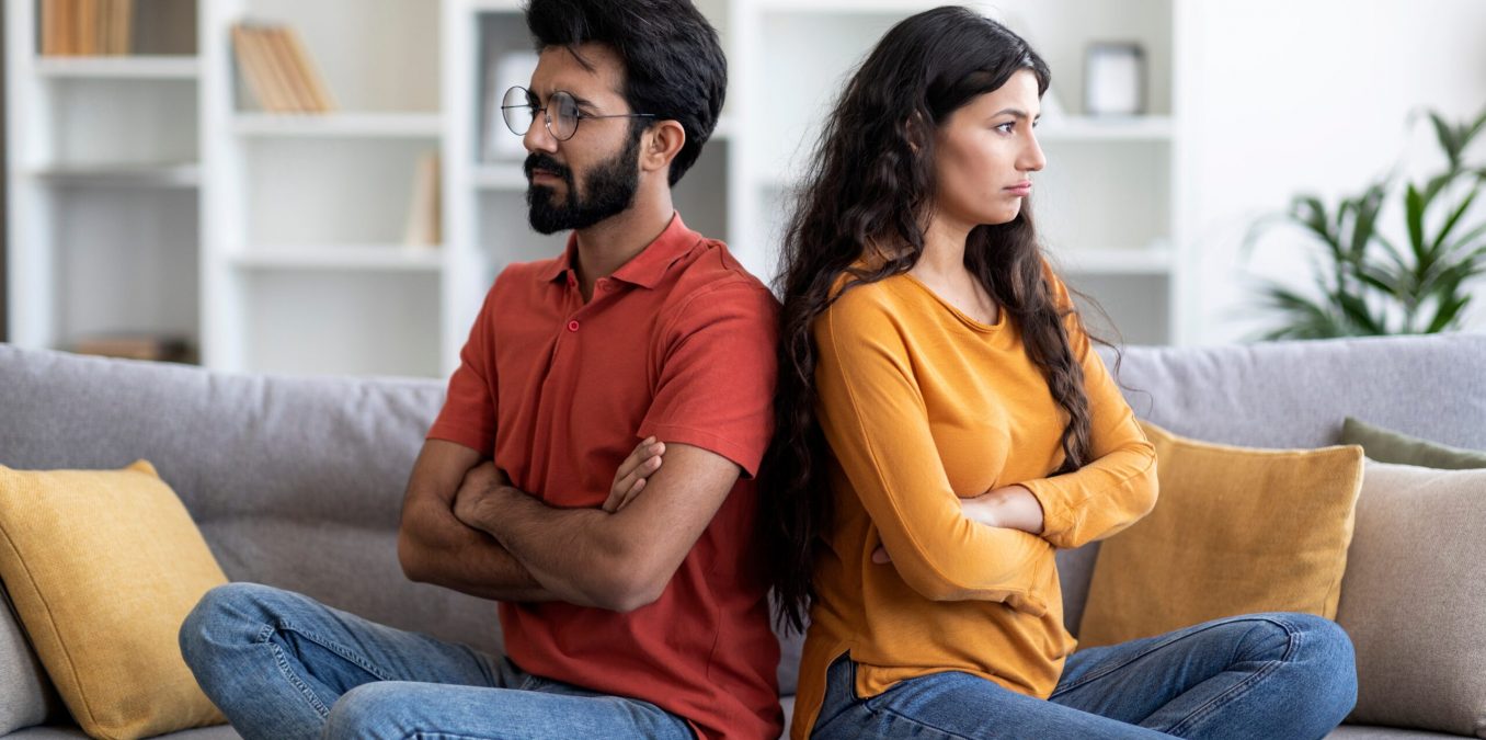 Young,Indian,Couple,Sitting,Back,To,Back,On,Couch,At