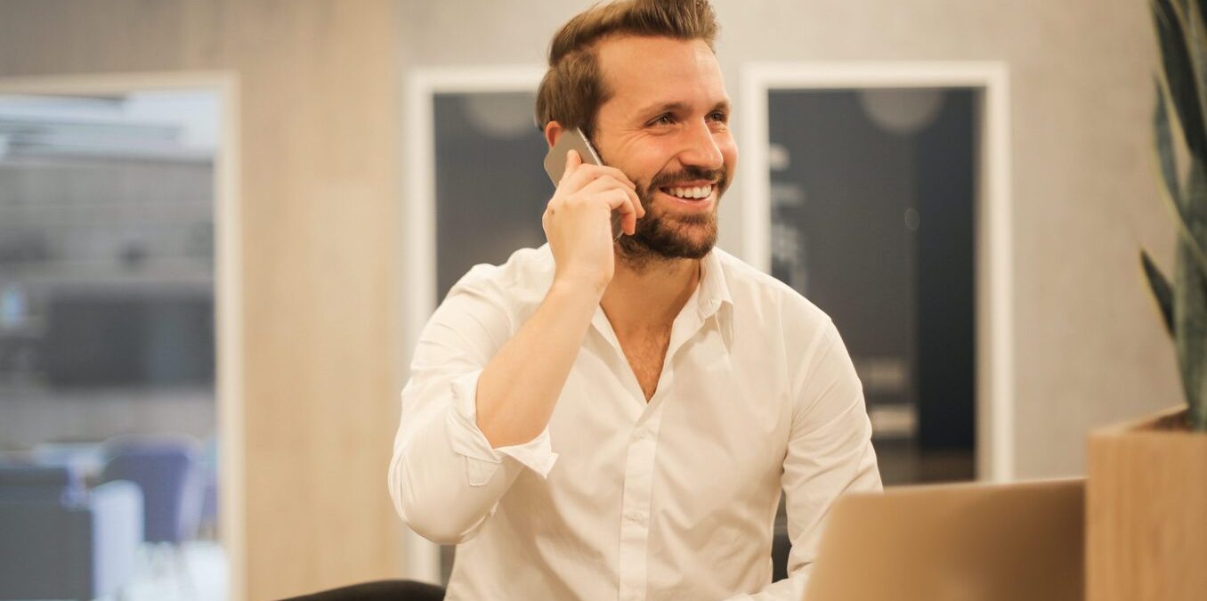 A smiling man with a beard and a white shirt sitting at a desk, talking on a smartphone in a modern office environment