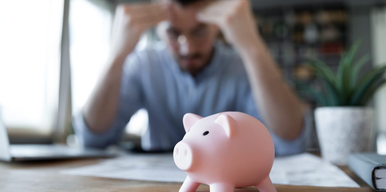 Stressed young male professional holding his head while looking at documents. Piggy bank in front