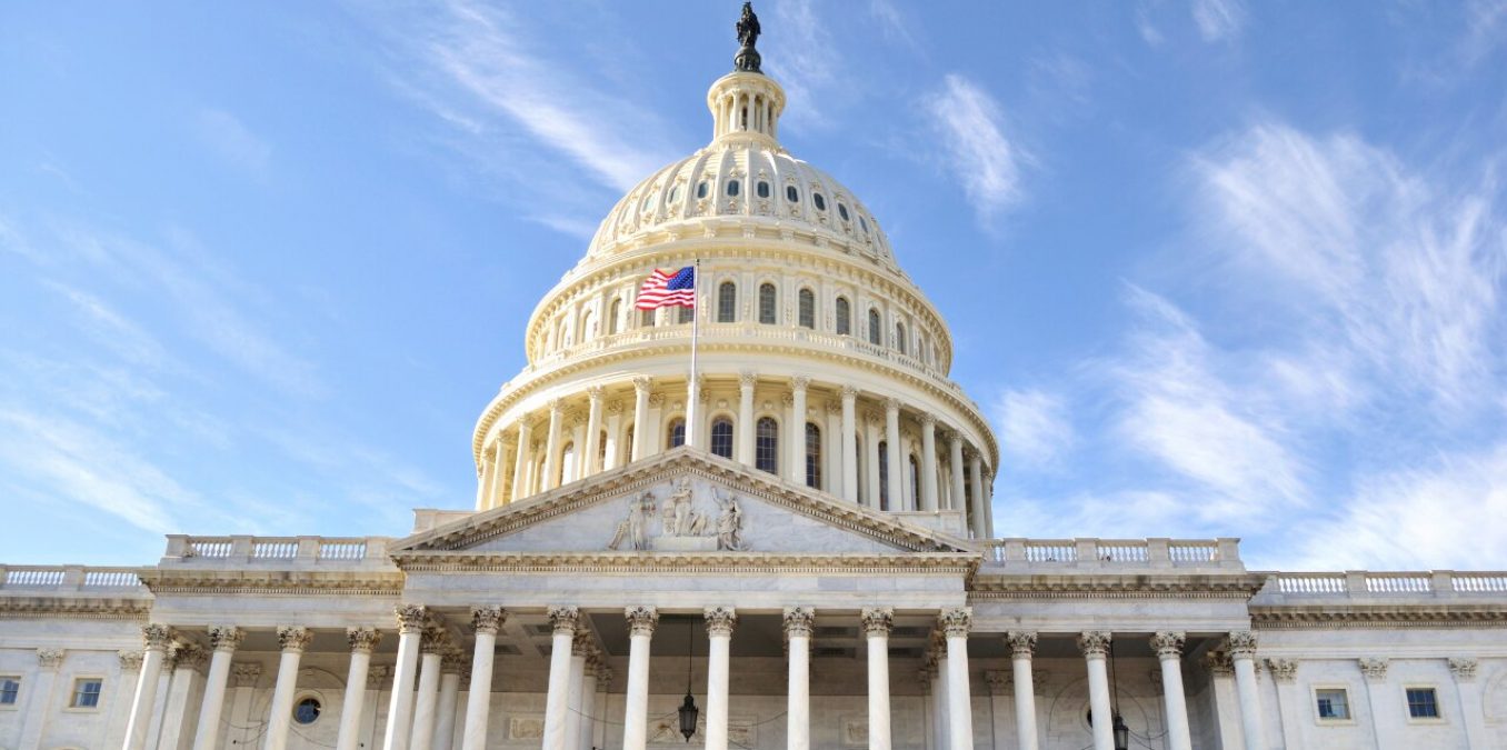 A low-angle shot of the United States Capitol Building in Washington, D.C., featuring the white dome and an American flag waving against a bright blue sky with wispy clouds.