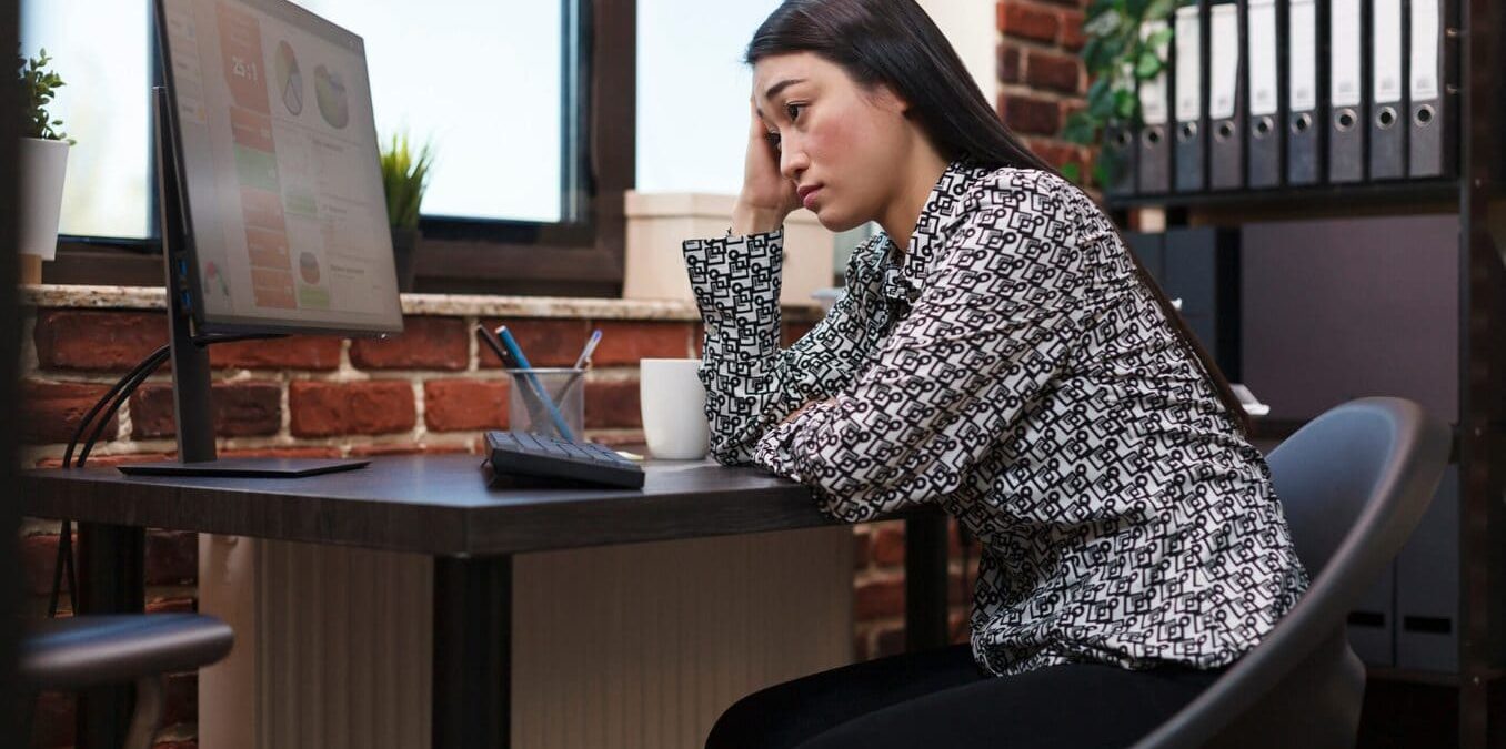 A woman sitting at a desk in a home office, looking stressed and resting her head on her hand while staring at financial charts on a computer monitor