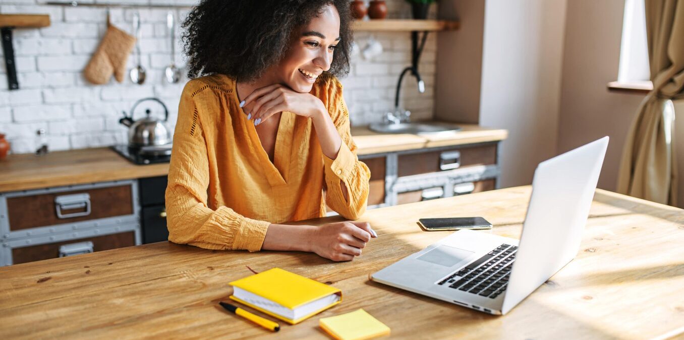 A smiling young woman with an afro hairstyle sitting at a light wooden kitchen table, looking at her laptop. She is wearing a mustard-yellow blouse and has a yellow notebook, pen, and smartphone on the table in front of her. The background shows a modern, rustic-style kitchen with white brick walls.