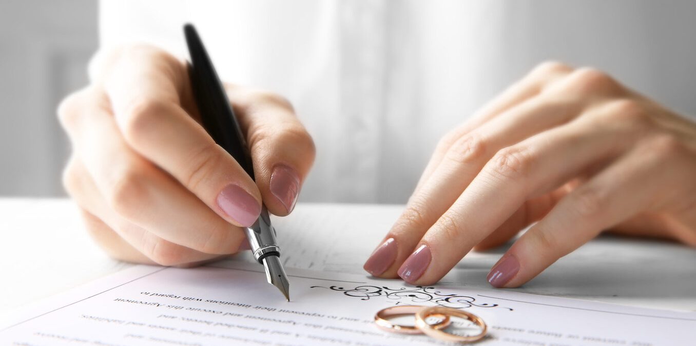 Close-up of a person signing a marriage certificate with wedding rings nearby