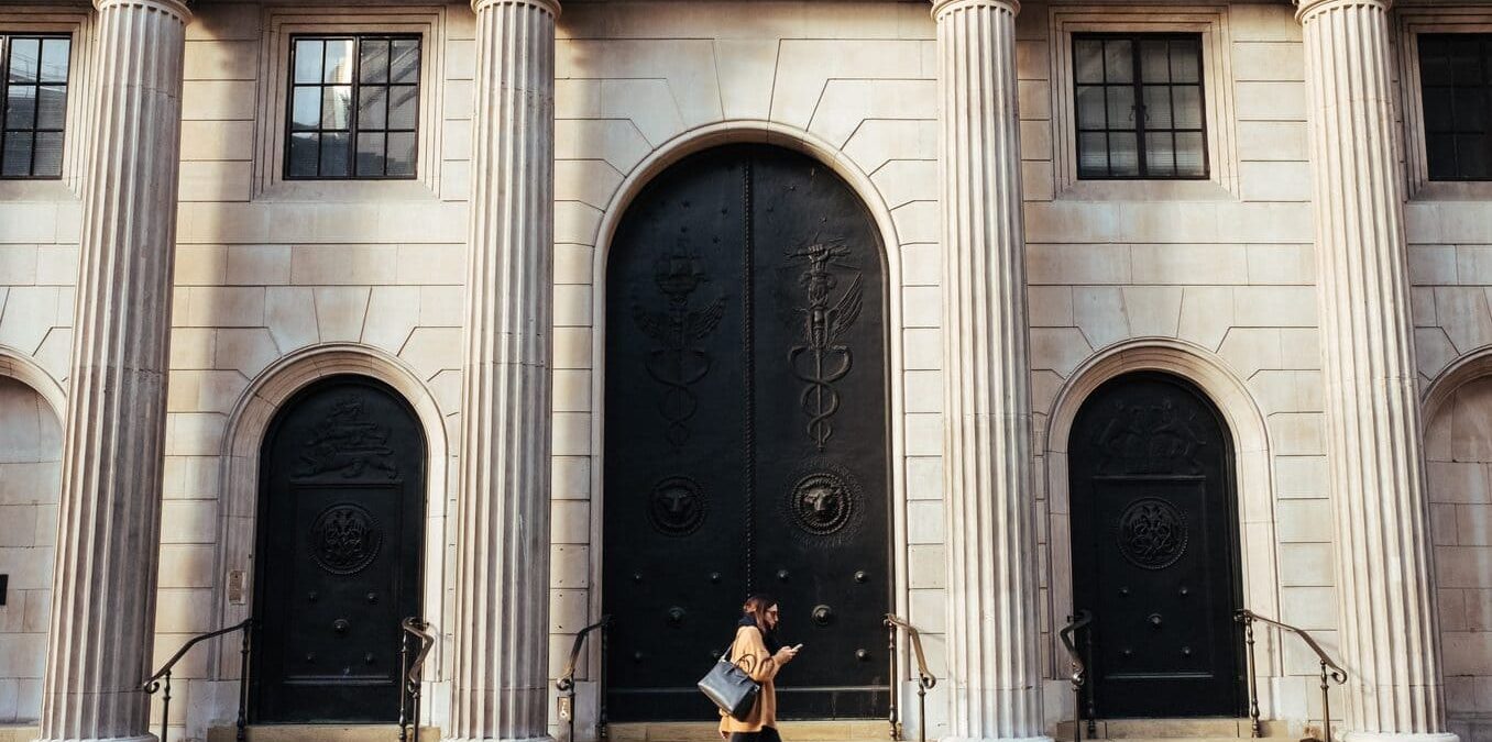 A woman walking along a sidewalk past a large stone building with tall Corinthian columns and ornate black arched doors