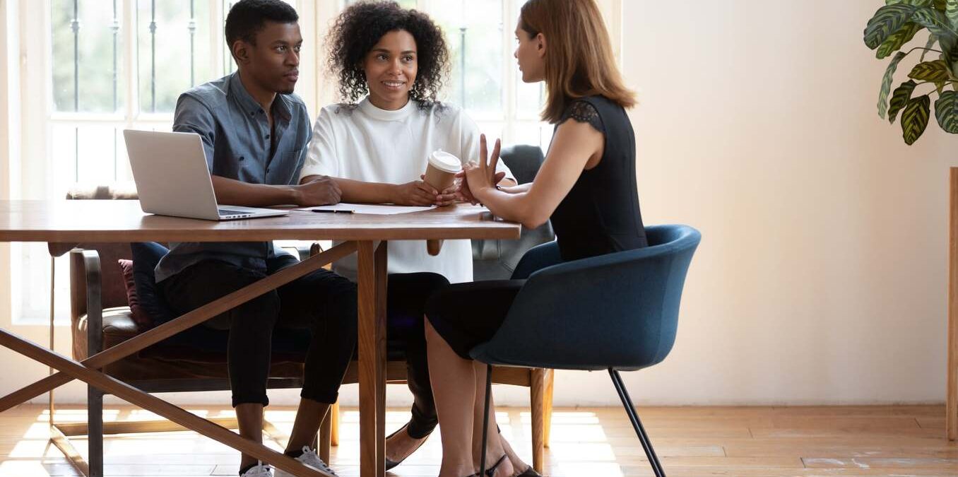 Young African American husband and wife sit at desk talk with female real estate agent discuss buying house together, biracial couple meet with designer architect or broker consult in modern office