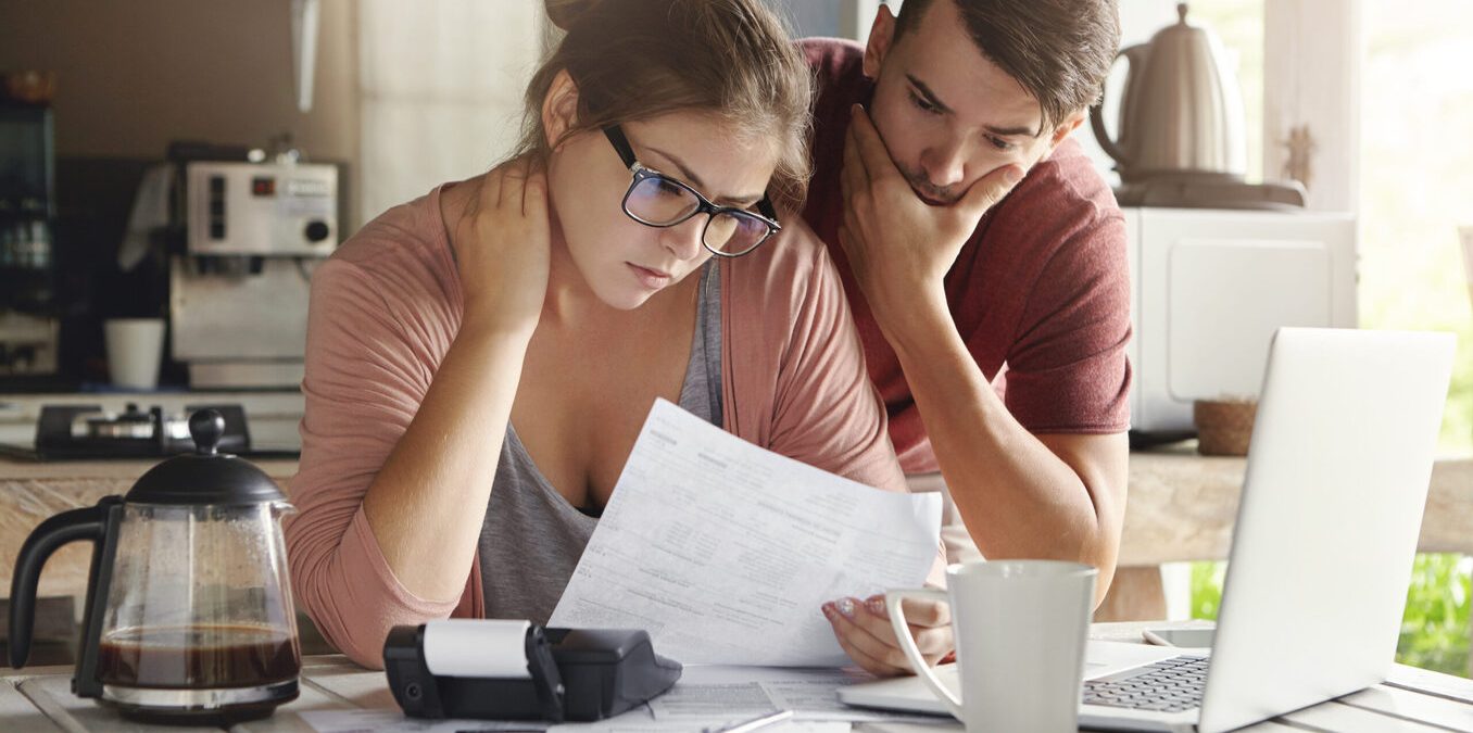 Young Caucasian family having debt problems, not able to pay out their loan. Female in glasses and brunette man studying paper form bank while managing domestic budget together in kitchen interior