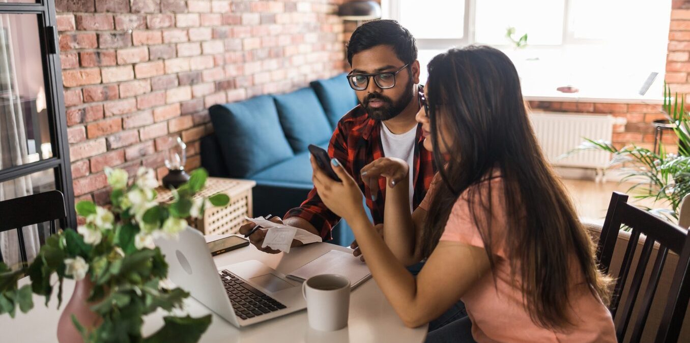 Young millennial indian husband and wife doing domestic paperwork, accounting job and reviewing paper bills, receipts at laptop computer, using online calculator and paying mortgage rent fees on